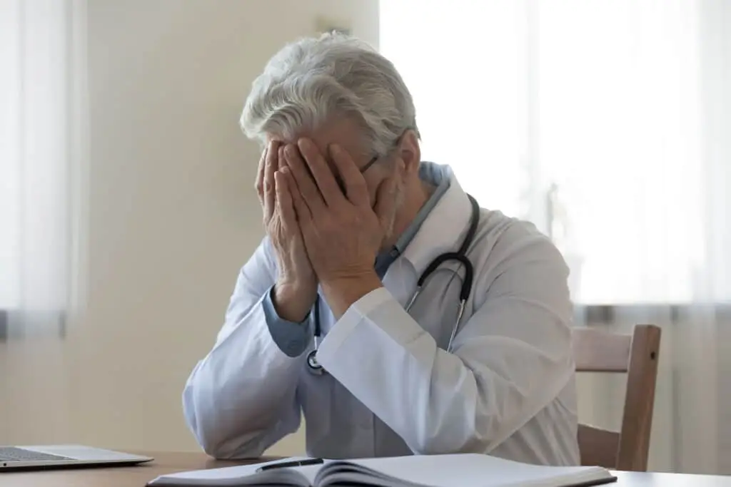 Stressed frustrated mature doctor wearing white uniform with stethoscope covering face with hands