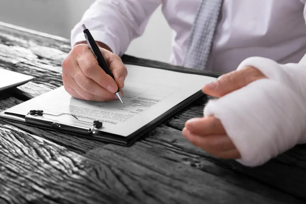 Close-up of a businessperson with broken arm filling health insurance claim form on wooden desk