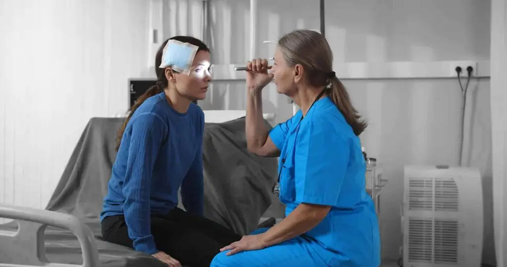Injured woman sitting on hospital bed with nurse woman shining in her eyes with flashlight.